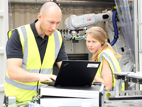 Two people operating a PC in front of a shipping container with a ABB robot